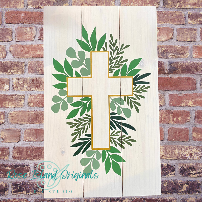 Decorative cross with green leaves on a wooden panel against a brick wall.