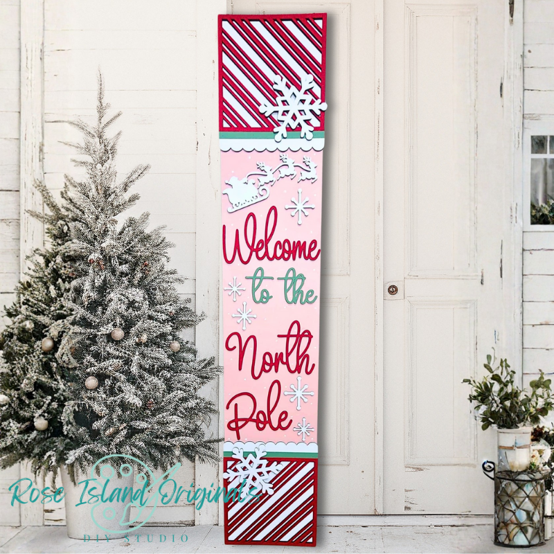 Decorative sign with 'Welcome to the North Pole' text in a festive design, placed against a white wooden door with a Christmas tree.