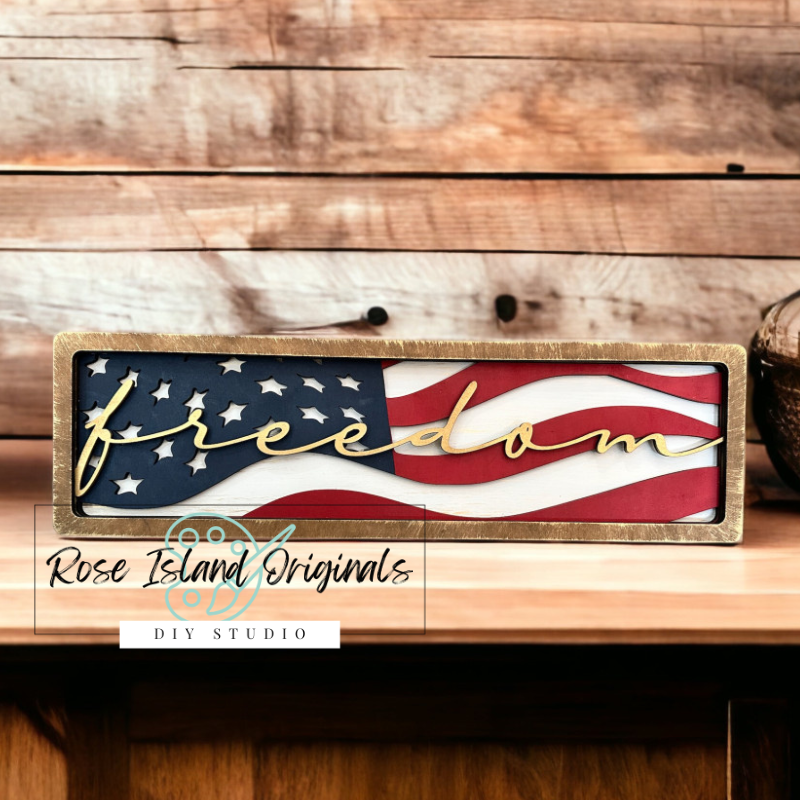 Wooden sign with American flag design and 'freedom' text on a wooden surface, featuring Rose Island Originals branding.