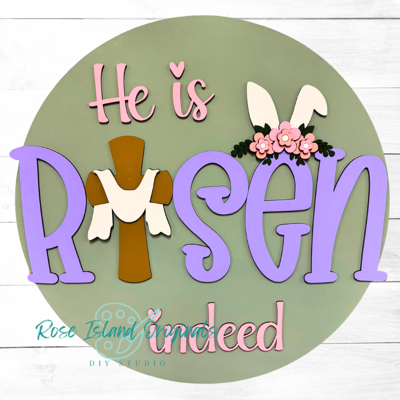 Decorative sign with 'He is Risen indeed' text, cross, and bunny ears on a green background.