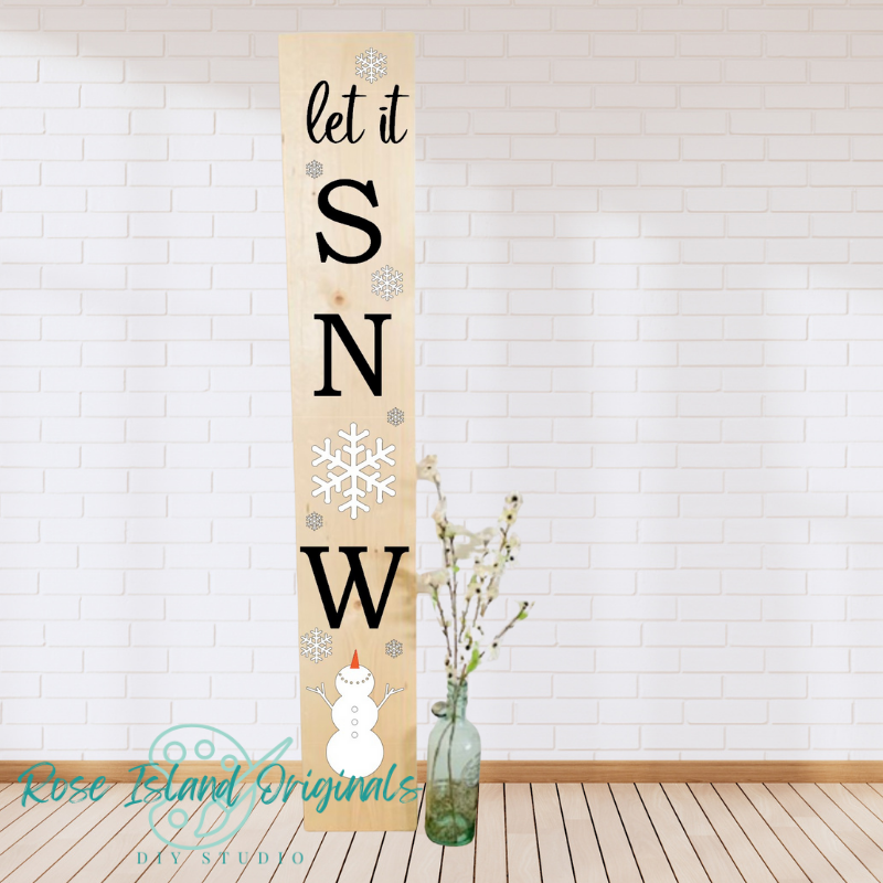 Decorative sign with 'let it snow' text and snowflake design on a wooden board, against a white brick wall.