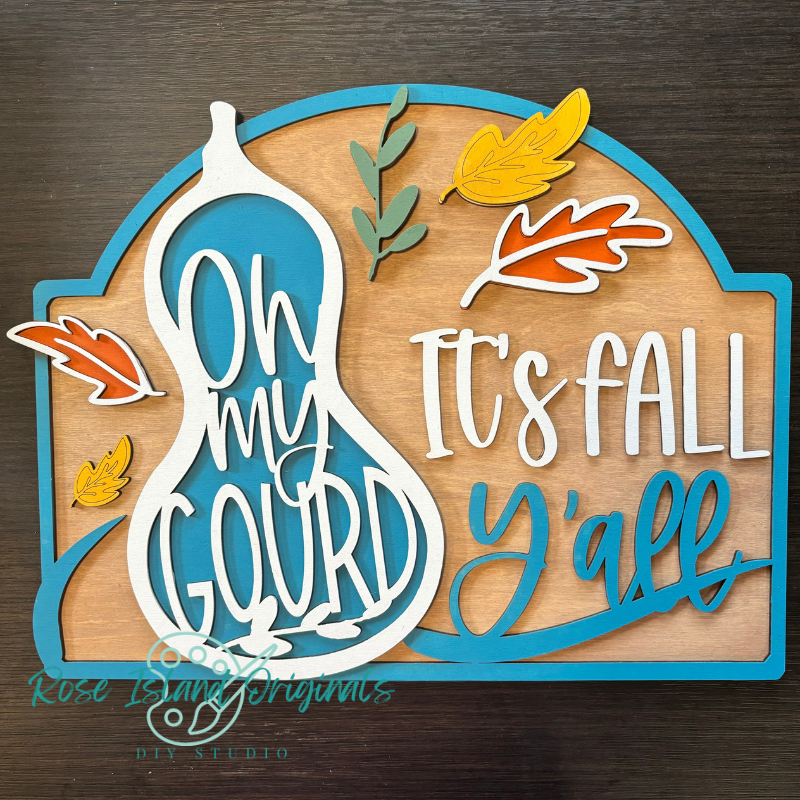 Decorative sign with 'Oh my gourd, it's fall y'all' text and autumn leaves on a wooden background.