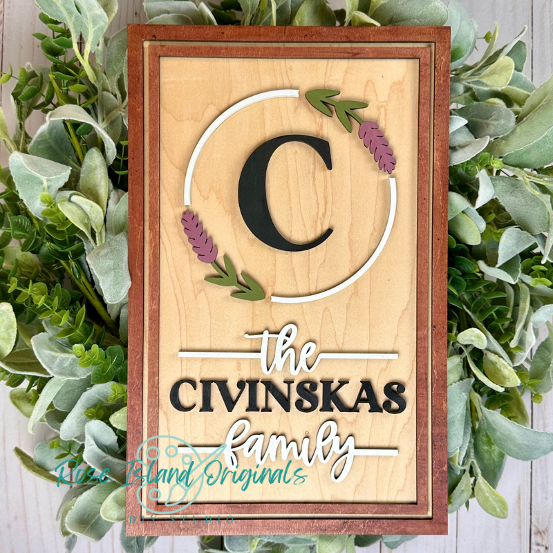 A personalized wooden sign with a light natural wood background, featuring a large black letter 'C' inside a white circular outline accented with green and purple leaves. Below, the text reads 'the CIVINSKAS family' in black and white script.