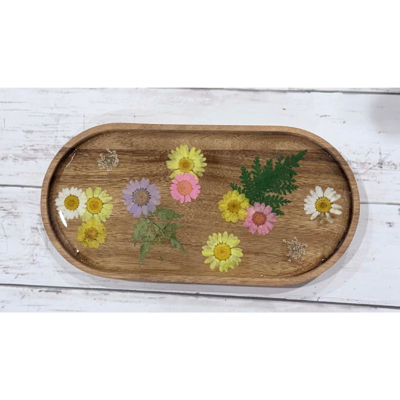 An oval wooden tray with a natural wood finish, decorated with pressed flowers and green leaves, displayed on a white-painted wooden surface.