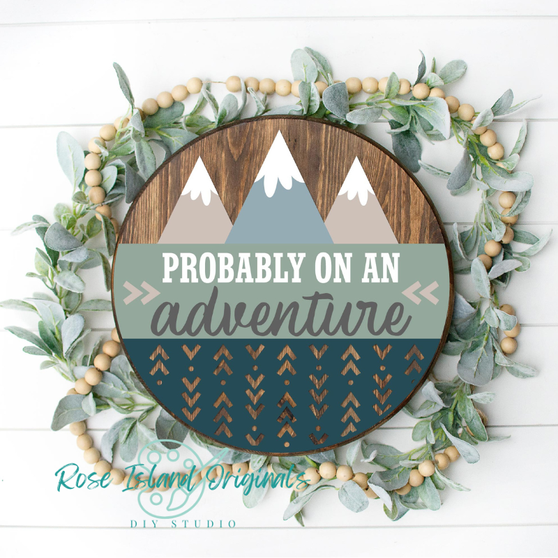 Decorative sign with mountains and text 'Probably on an adventure' surrounded by greenery and wooden beads.