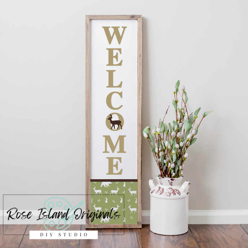 Decorative 'WELCOME' sign with gold text and wooden frame on a white wall, next to a white vase with greenery.