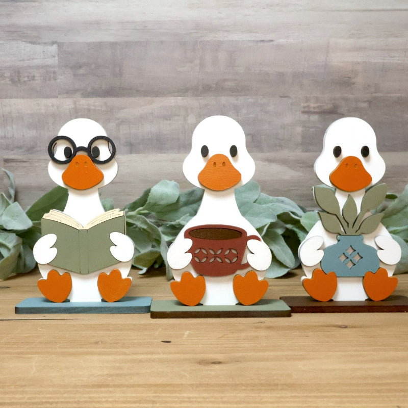 Three handcrafted wooden duck figurines sitting on a wooden surface with green leaves behind them. The left duck wears black glasses and holds an open book.