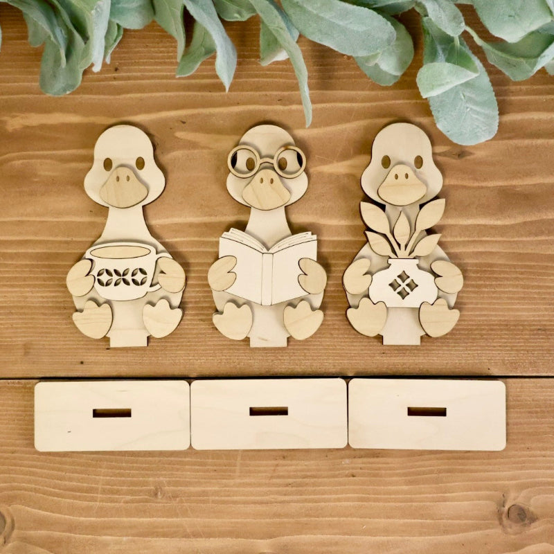 Three laser-cut wooden goose figurines arranged on a rustic wooden surface, with three matching rectangular wooden boxes below them. The ducks are styled with green foliage at the top.