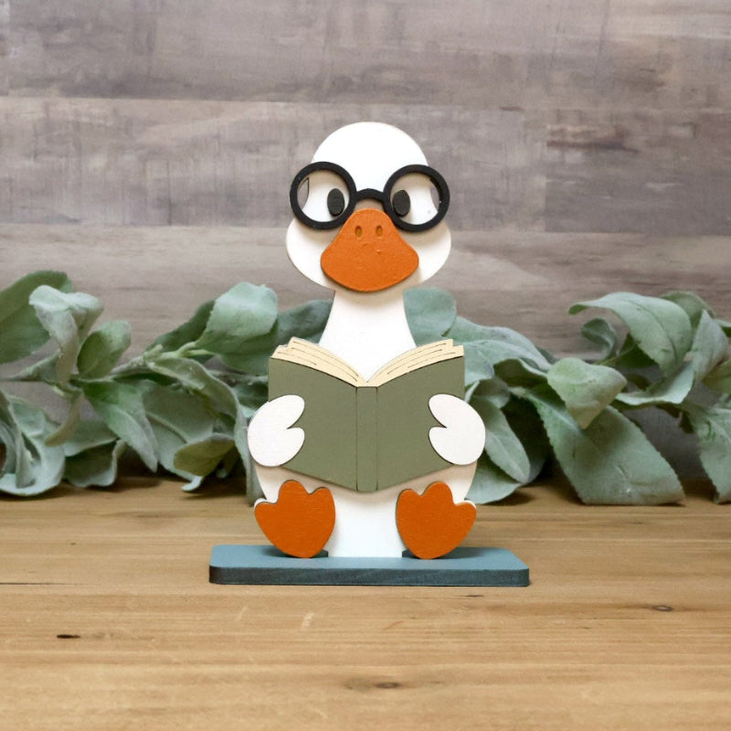 A wooden duck figurine wearing black glasses and holding an open book, sitting on a blue base on a wooden tabletop with green leaves and a wood plank wall background.