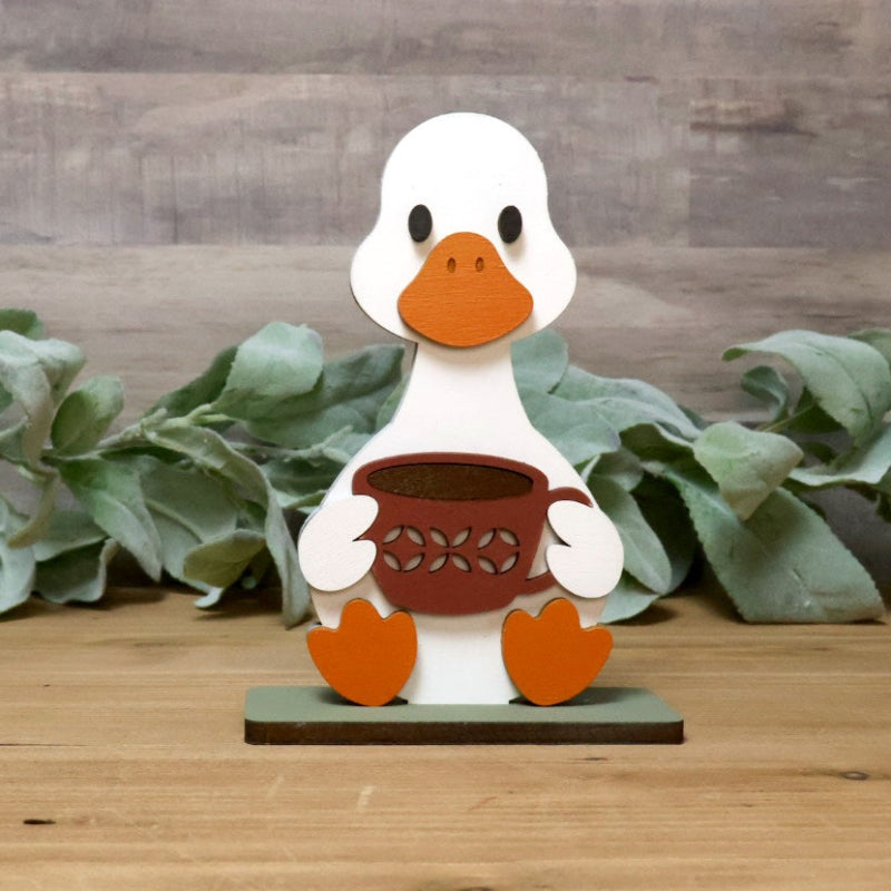 A small white duck figurine with an orange beak and feet, holding a brown basket, displayed on a wooden surface with green leaves and a wood plank background.