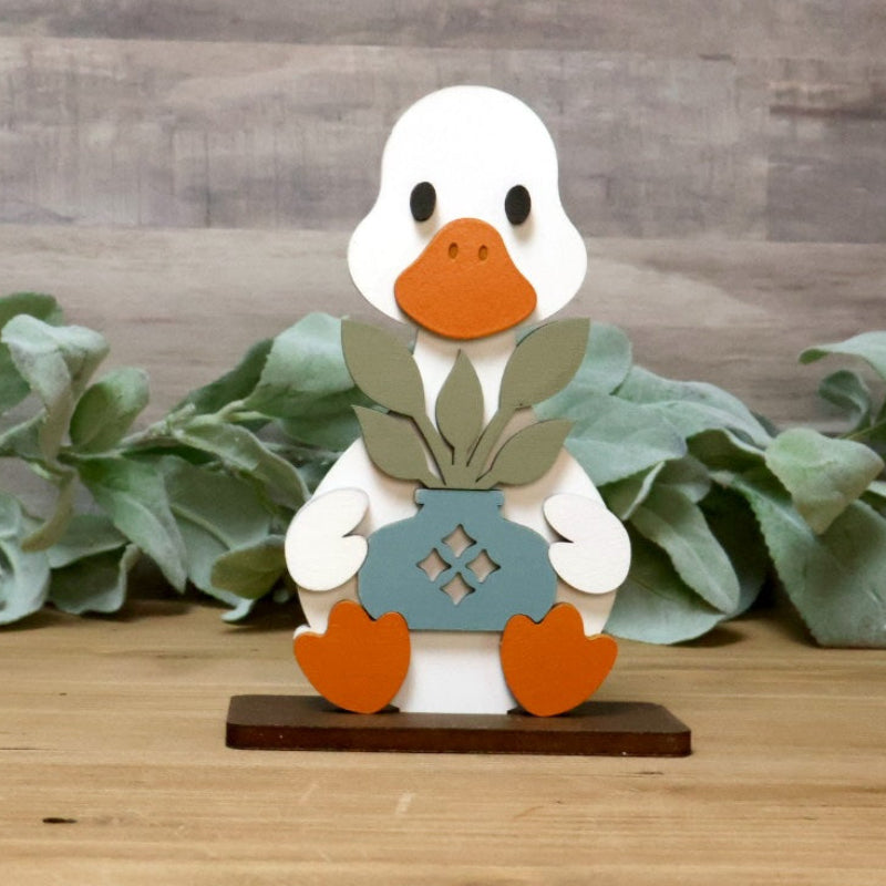 A small decorative goose figurine stands centered on a wooden surface against a rustic wood plank backdrop, with a green leaf garland running behind it. The goose is white with an orange beak and orange feet, wearing a gray vest and holding a blue-gray box.