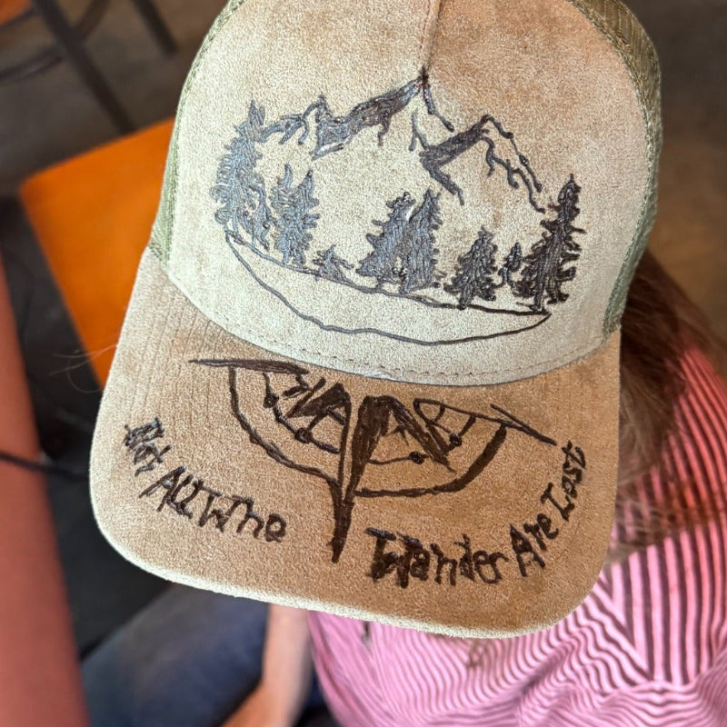A person seated at a table wearing a distressed tan trucker hat with a mountain and tree graphic on the front and handwritten text on the brim, photographed in a casual indoor setting.