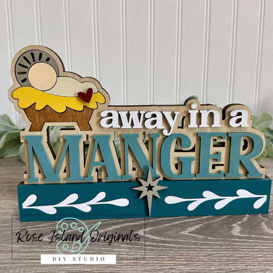 Decorative sign with 'away in a MANGER' text and Christmas scene elements on a wooden surface.