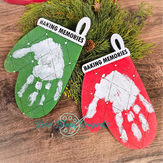 Two Christmas ornaments shaped like green mittens and red oven mitts with handprints, labeled 'Baking Memories', on a wooden surface with greenery.
