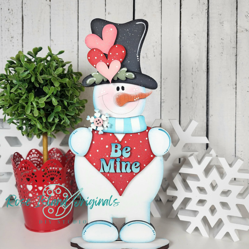 Decorative snowman with heart and 'Be Mine' sign, surrounded by festive decor on a wooden background.
