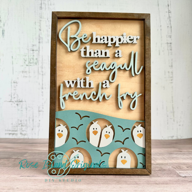 A framed wooden sign with layered cutout text reading 'Be happier than a seagull with a french fry' in white and pale blue, plus a teal band at the bottom featuring cartoon penguins and black birds. The sign is photographed standing upright on a wood surface against a whitewashed wall.