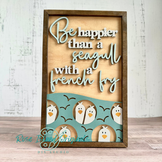 A framed wooden sign with layered cutout text reading 'Be happier than a seagull with a french fry' in white and pale blue, plus a teal band at the bottom featuring cartoon penguins and black birds. The sign is photographed standing upright on a wood surface against a whitewashed wall.