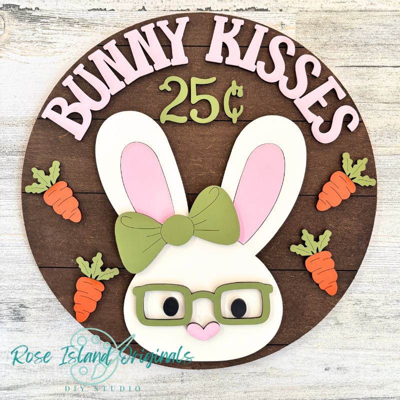 Decorative sign with a bunny face, carrots, and 'Bunny Kisses 25 cents' text on a wooden background.