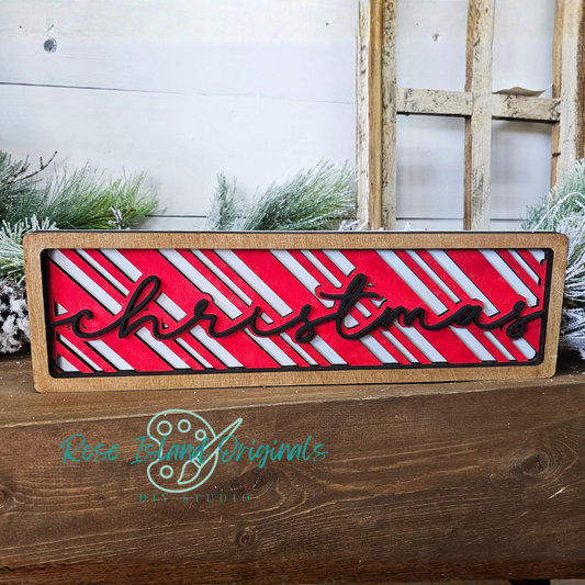 Decorative sign with 'Christmas' text on a red and white striped background, placed on a wooden surface with a rustic setting.