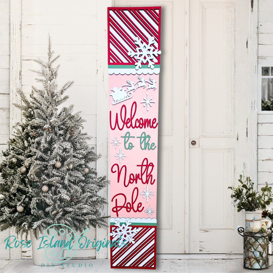 Decorative sign with 'Welcome to the North Pole' text in a festive design, placed against a white wooden door with a Christmas tree.
