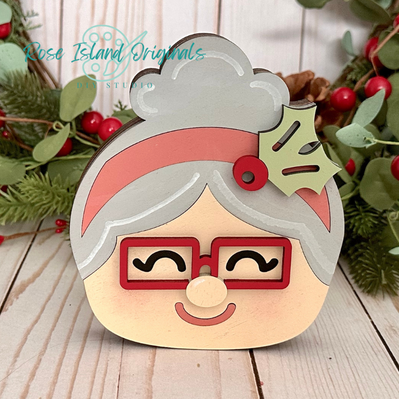 Wooden ornament with a Santa Claus face design, featuring a red hat, glasses, and gray hair, on a wooden surface with greenery.