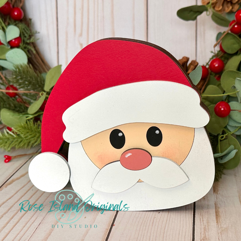 Cartoon Santa Claus face cutout with a red hat and white beard on a wooden surface with Christmas decorations.