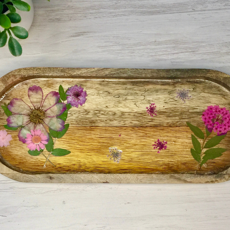 A rustic rectangular wooden tray with rounded corners, decorated with pressed flowers and green leaves, displayed on a light gray wood-textured surface.
