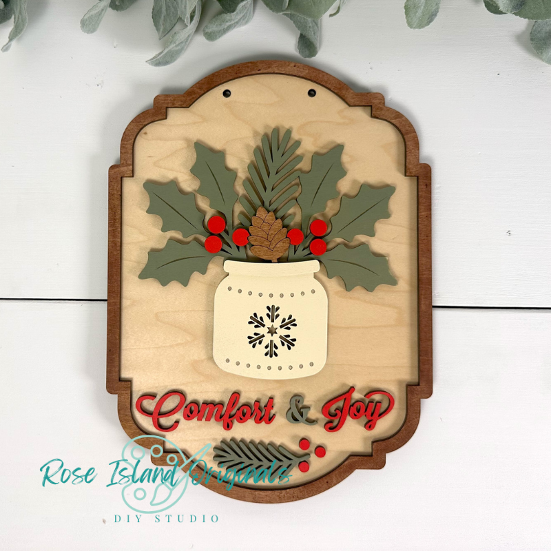 Decorative wooden sign with holly leaves, berries, and a snowflake design, featuring the text 'Comfort & Joy'.