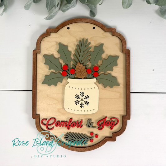 Decorative wooden sign with holly leaves, berries, and a snowflake design, featuring the text 'Comfort & Joy'.