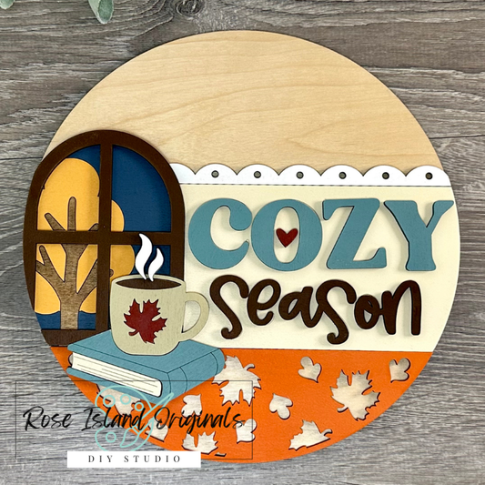 Decorative sign with 'Cozy Season' text, cup of coffee, book, and autumn leaves on a wooden background.