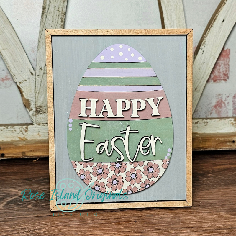 Framed decorative sign with 'Happy Easter' text inside a stylized egg on a wooden surface.