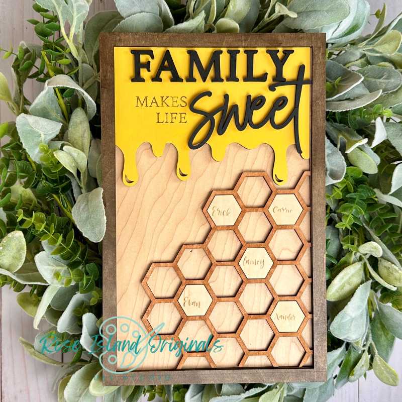 A rectangular wooden sign with a honeycomb design and the words “FAMILY makes life sweet” in yellow and black, styled with green foliage around it. The honeycomb cells are labeled with words like “Love,” “Honey,” and “Flavor.