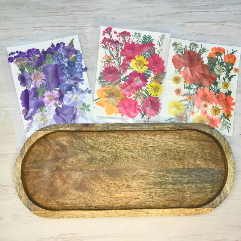 A rustic oval wooden tray with a raised rim sits on a light wood surface, with three colorful pressed flower cards arranged behind it.