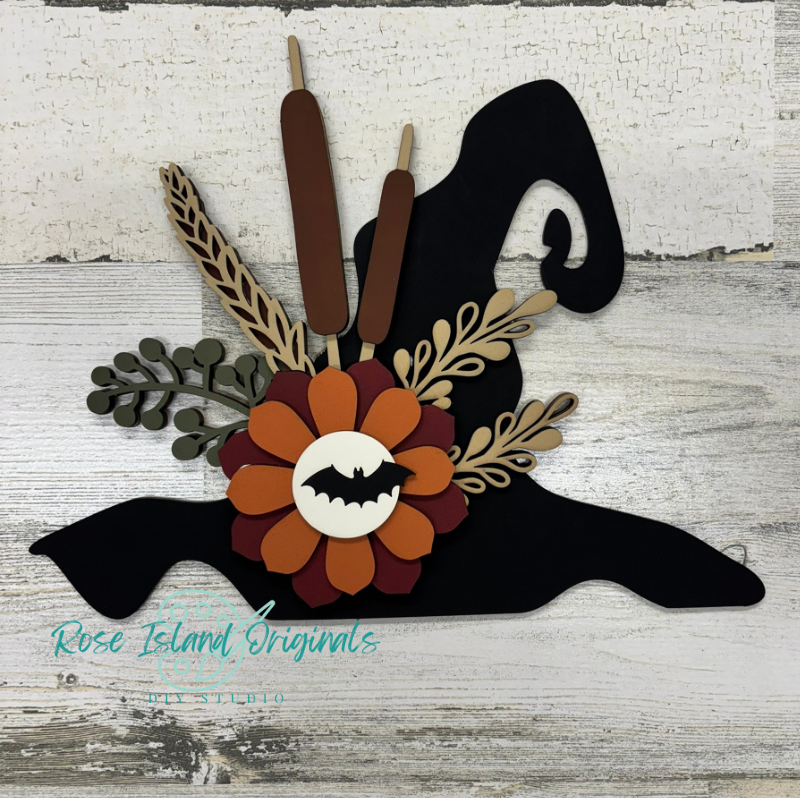 A decorative layered paper cutout of a witch hat with autumn and Halloween elements, including a flower with a bat in the center, two candles, leaves, and cattails, displayed on a distressed white wood background. A watermark for 'Rose Island Originals' is visible in the lower left.