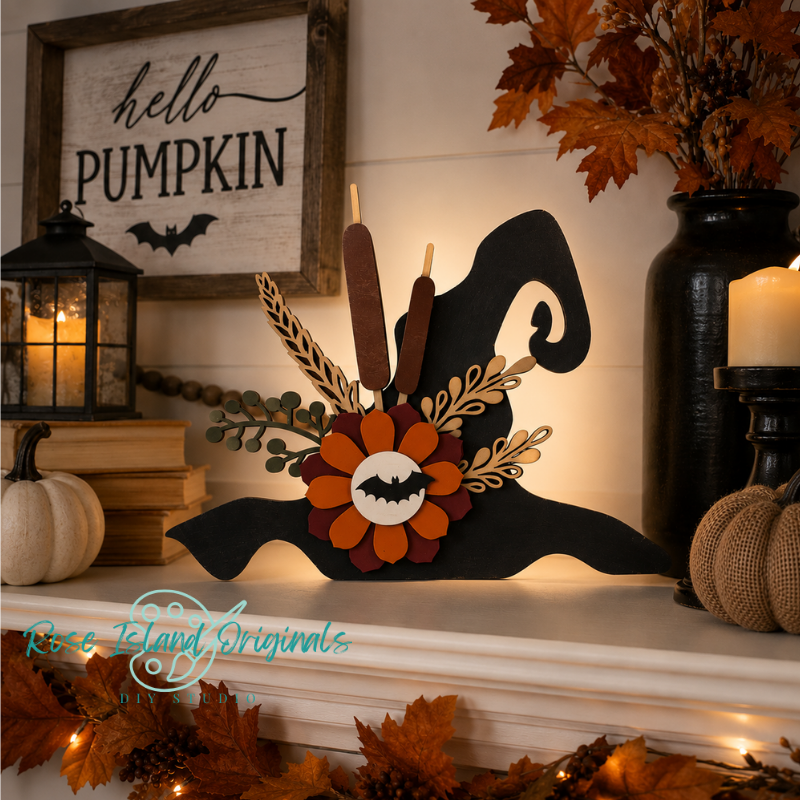 A Halloween-themed decorative candle holder shaped like a witch hat, displayed on a mantel with autumn decor. The black silhouette features a layered orange and brown flower with a bat motif, dried leaves, and two brown candles.