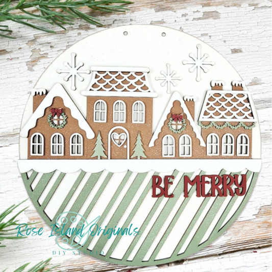 Decorative wood cutout with gingerbread house design and 'Be Merry' text on a wooden surface.