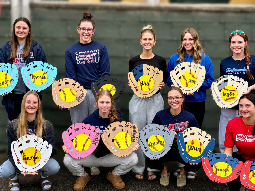 A group of 14 female softball players posing together outdoors, each holding a large decorative softball glove prop with a softball graphic and personalized names like 'Meadows', 'Harris', and 'Pry'.