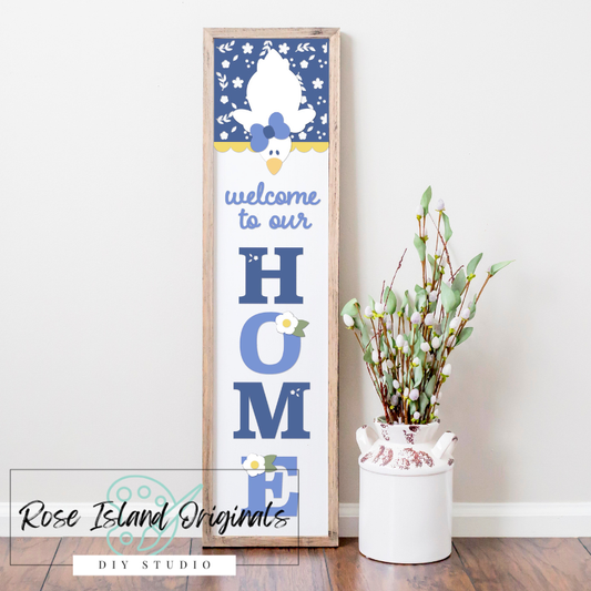 Decorative sign with 'welcome to our HOME' text and a goose design, placed on a wooden floor with a vase of flowers.