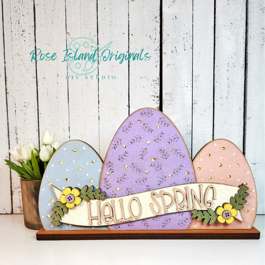 Decorative sign with colorful eggs and 'Hello Spring' text on a wooden stand, against a white wooden panel background.
