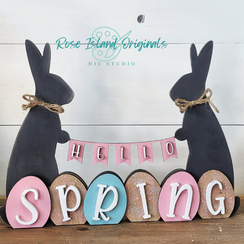 Decorative sign with bunnies and 'Hello Spring' text on a wooden surface, featuring Rose Island Originals branding.