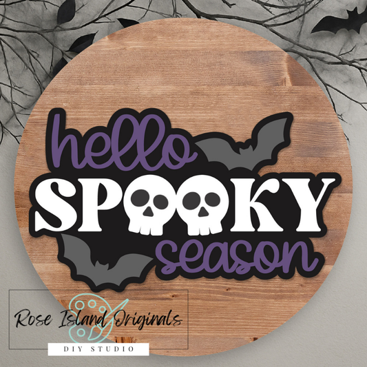 Wooden round sign with 'hello spooky season' text with skulls and bats on a Halloween-themed background.