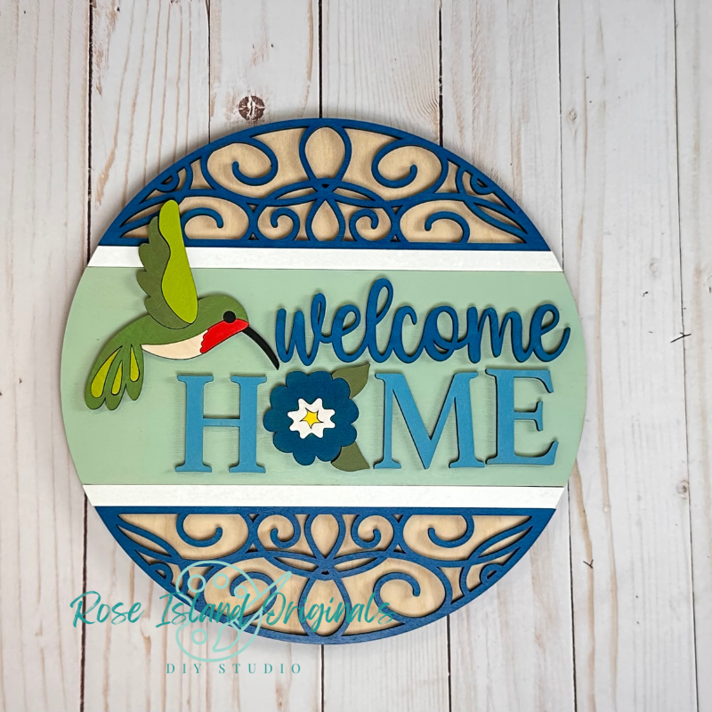 Decorative sign with 'Welcome Home' text and a hummingbird design on a wooden background.