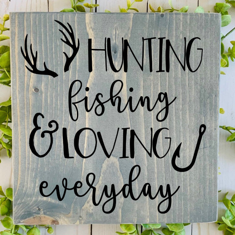 Wooden sign with 'Hunting fishing & loving everyday' text on a white wooden background with greenery.