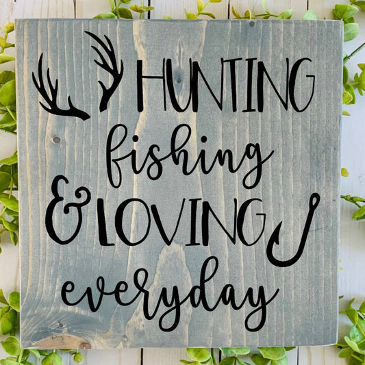Wooden sign with 'Hunting fishing & loving everyday' text on a white wooden background with greenery.