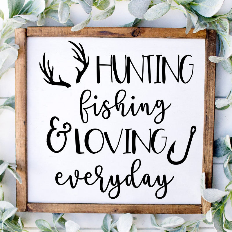 Wooden sign with 'Hunting fishing & loving everyday' text surrounded by greenery on a white background