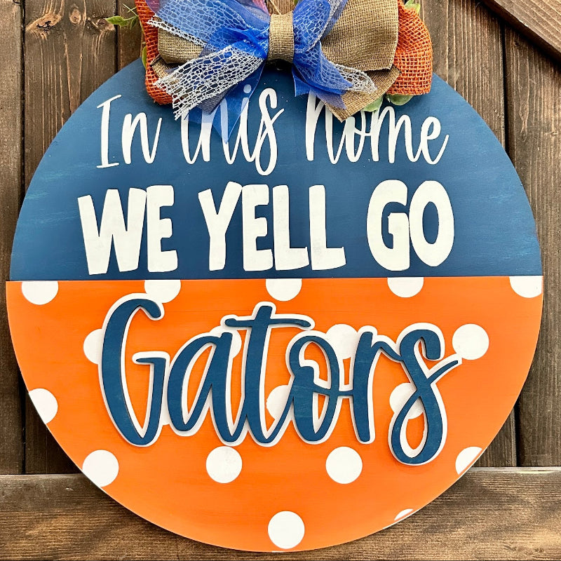 A round wooden fall wreath sign with a blue and orange color-blocked design, white polka dots, and the text 'In this home WE YELL GO Gators'. It features a decorative bow at the top with blue mesh, orange burlap, and tan burlap.