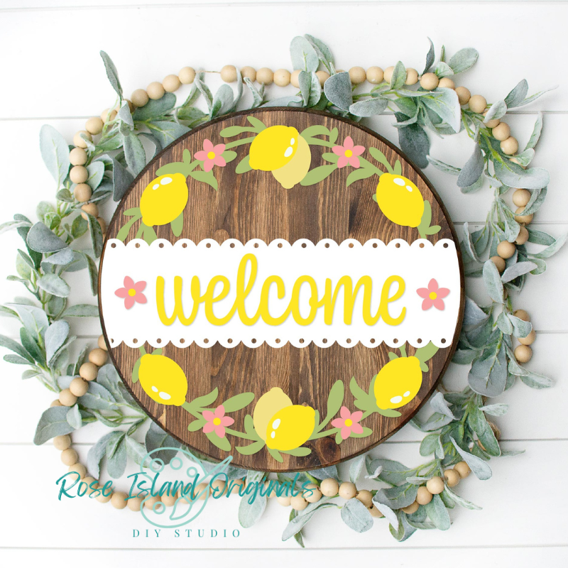 A circular wooden welcome sign with a white banner reading 'welcome' in yellow script, surrounded by illustrated yellow lemons, pink flowers, green leaves, and a wooden bead wreath on a white shiplap background. A watermark at the bottom reads Rose Island Creations DIY Studio.