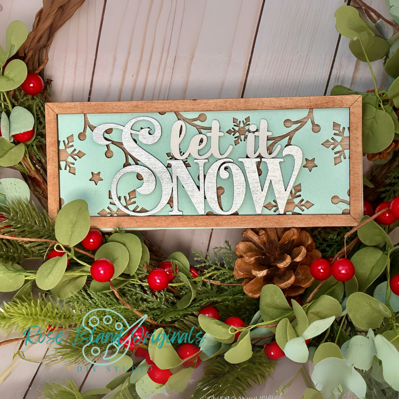 Decorative sign with 'Let it Snow' text surrounded by Christmas foliage on a wooden surface.