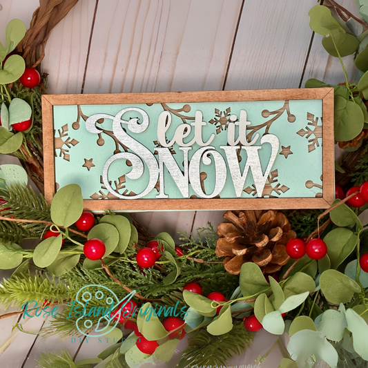 Decorative sign with 'Let it Snow' text surrounded by Christmas foliage on a wooden surface.
