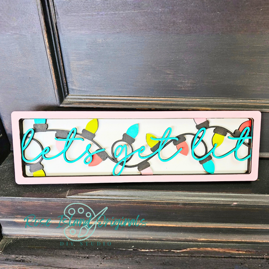 Decorative sign with 'let's get lit' text and Christmas lights design on a wooden surface.